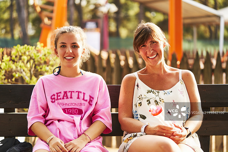 母女肖像，穿着休闲服装，坐在公园长椅上微笑着看向镜头，正面视角图片素材