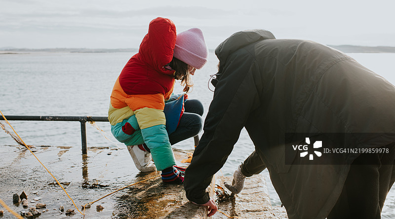 穿着五彩斑斓羽绒服的孩子蹲在码头上，成年人在阳光明媚、寒冷的天气中给她展示一只螃蟹。图片素材