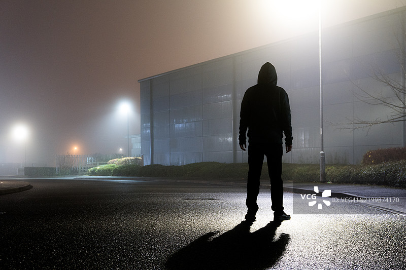 一个阴险的戴兜帽的人影，在神秘的雾夜中被街灯勾勒出轮廓，站在空旷的工业区。图片素材
