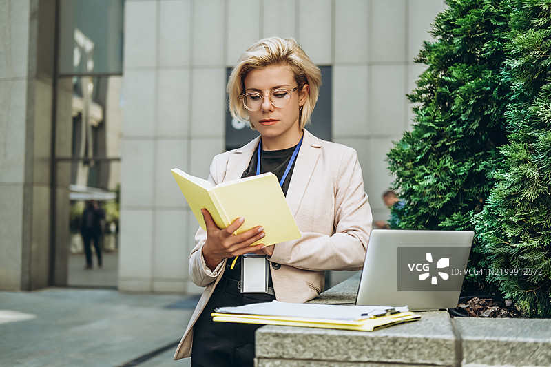 专注的年轻女性在办公中心附近的紫杉树旁阅读一本书，旁边有一台超薄笔记本电脑。图片素材