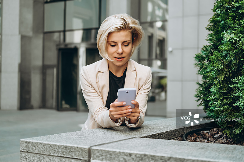 一位穿着浅色西装外套的愉快女性在商业中心附近的柏树旁使用智能手机。图片素材
