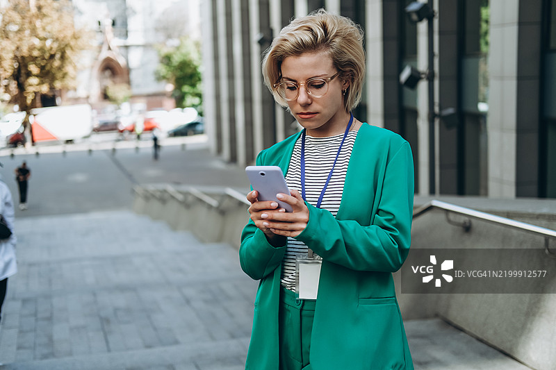戴眼镜的年轻女性穿着青绿色西装，在楼梯上使用智能手机。图片素材