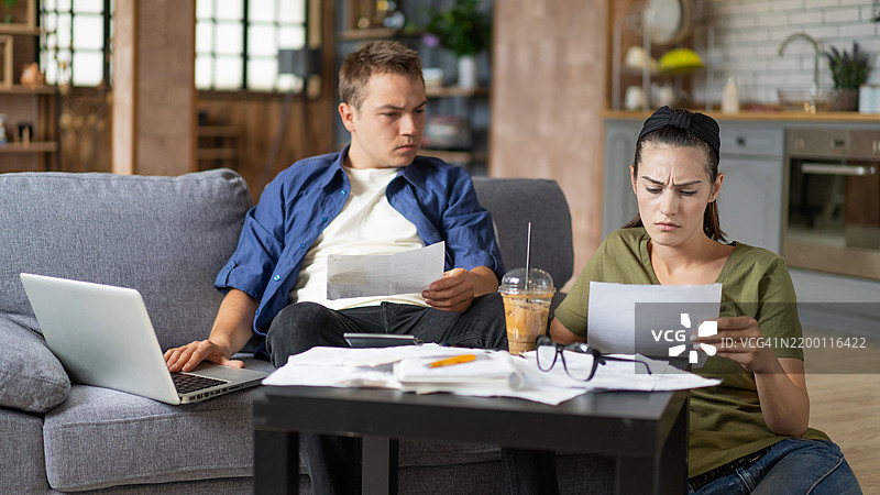 年轻夫妇在家中使用平板电脑和计算器计算账单。年轻夫妇坐在沙发上使用电脑计算财务。年轻男子与妻子在家中分析他们的财务文件。图片素材