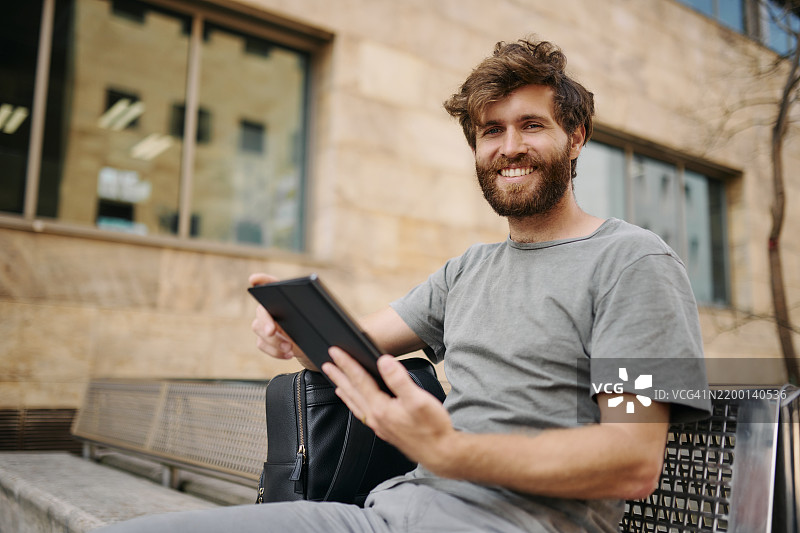 快乐的男人，坐在长椅上，手持平板电脑，用于旅行、导航或城市旅游应用的地图指引。微笑的男性，游客或旅行者，利用科技进行旅行、通勤或移民。图片素材