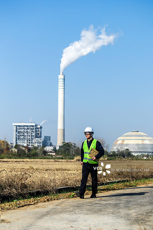 男性首席工程师穿着安全背心、硬帽和护目镜，在现代联合循环发电厂进行测量记录。图片素材
