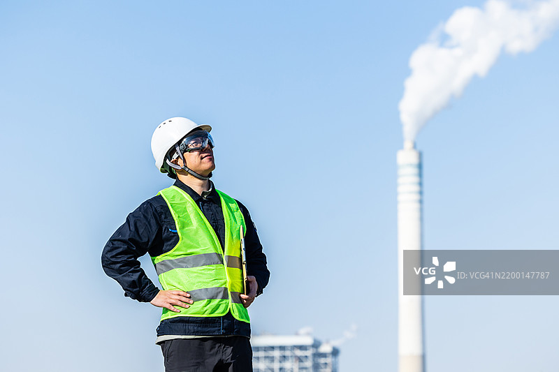 男性首席工程师穿着安全背心、硬帽和护目镜，在现代联合循环发电厂进行测量记录。图片素材