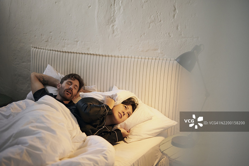 在卧室里睡觉、打鼾的情侣，女人在床上因噪音感到沮丧和失眠。关系紧张，因声音而烦恼，男人在夜里吵闹，女人捂住耳朵，疲惫不堪。图片素材