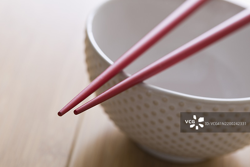 红色筷子放在一个带有纹理设计的白色碗上，桌子是木质的。图片素材