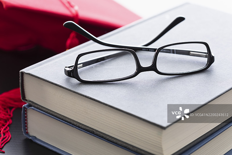 毕业庆祝活动中，桌子上放着书籍和眼镜，背景有一顶学位帽。图片素材