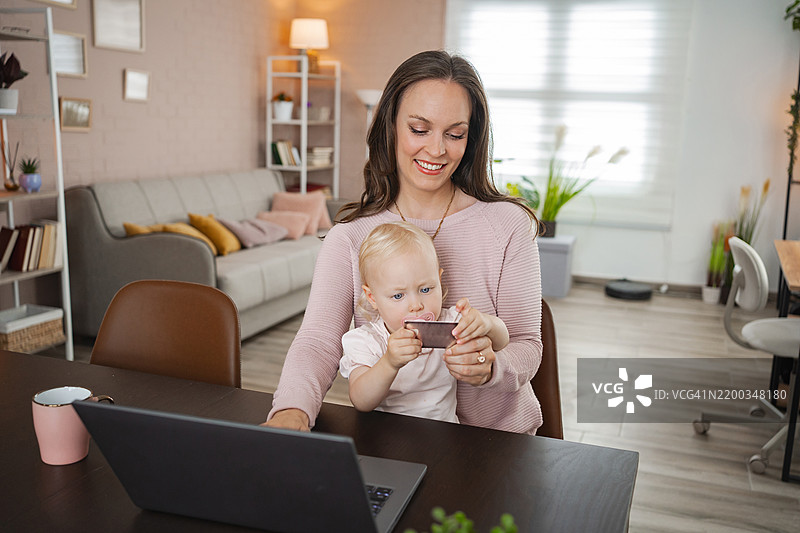 母亲在家工作，同时抱着她的女儿，女儿正在玩一张信用卡。图片素材