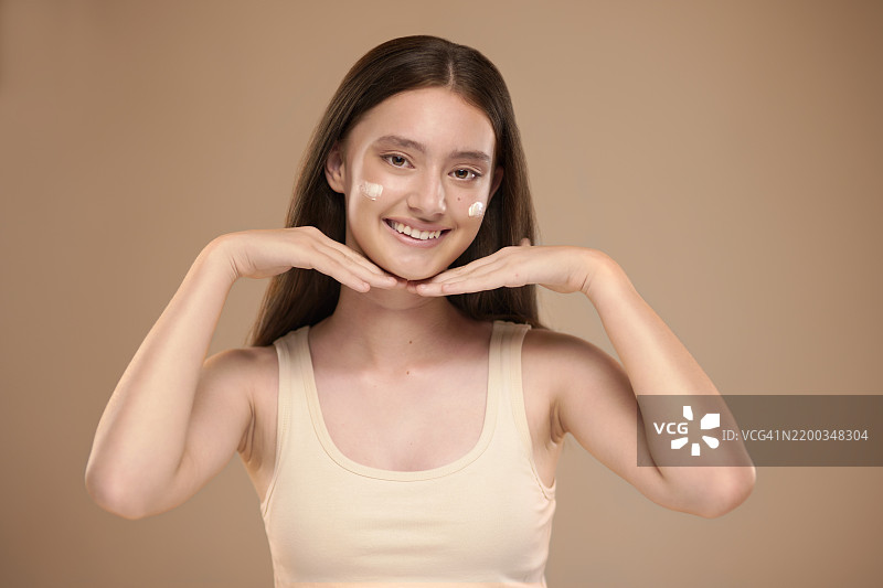 女孩在工作室肖像中微笑，骄傲地使用化妆品和面霜，背景中有保湿霜的应用。青少年，快乐的模特，使用护肤霜、精华或美容产品。图片素材