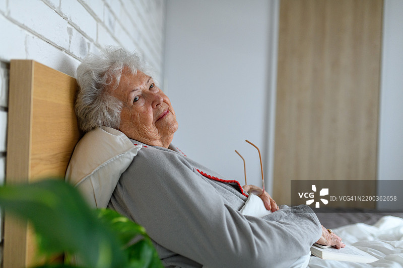 一位老年女士躺在床上，手中拿着一本好书。图片素材