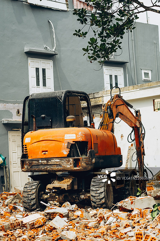 一台橙色挖掘机停在建筑工地上，周围堆满了破碎的砖块和 debris。一名穿黑色衣服的工人站在附近，监督着正在进行的拆除工作。图片素材
