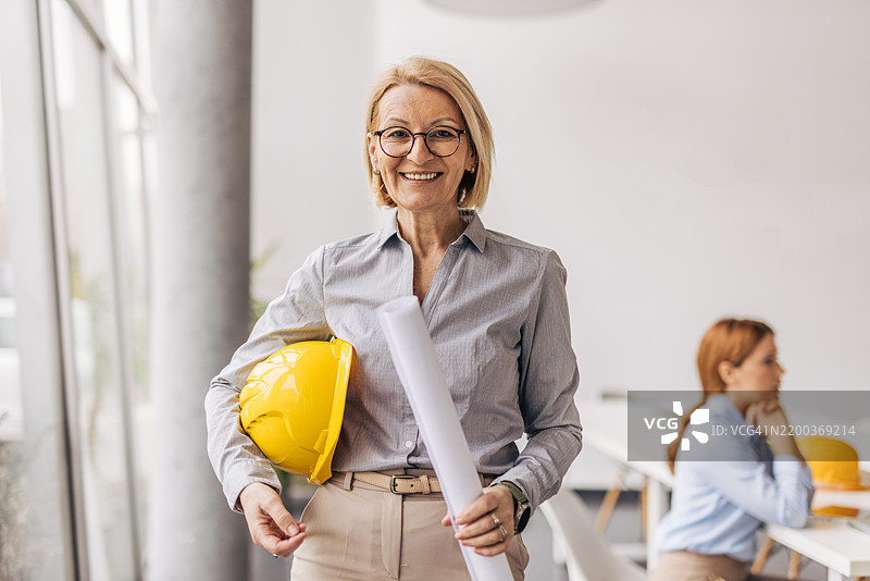 微笑着看向镜头的资深女性建筑师，手持蓝图和安全帽图片素材