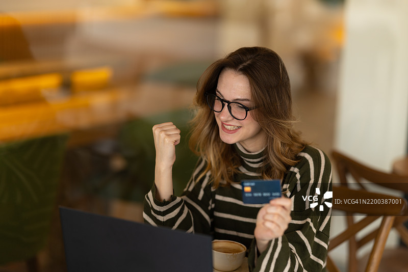 兴奋的年轻女性在咖啡馆使用信用卡和笔记本电脑进行在线支付，庆祝成功购买。图片素材