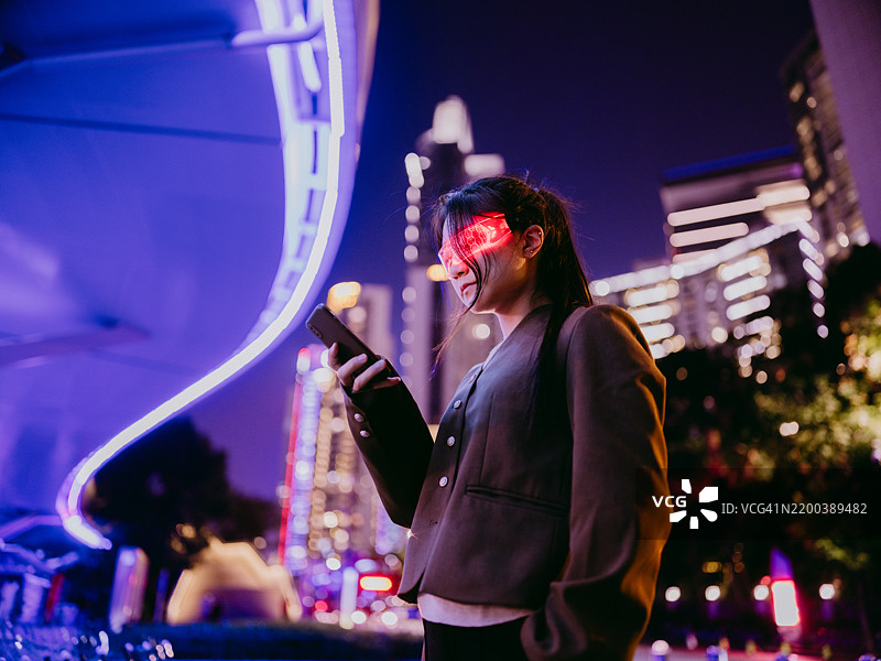 自信的女商人佩戴虚拟现实眼镜，手持智能手机，站在发光的人行天桥下图片素材