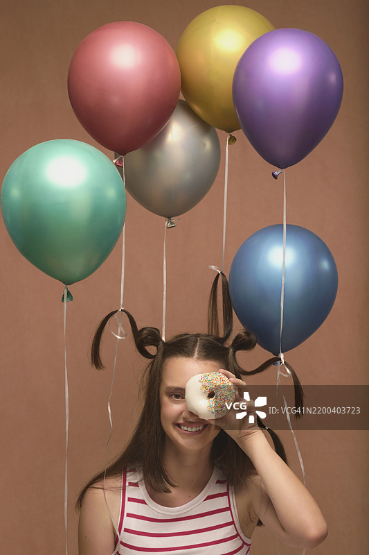女孩、甜甜圈和气球在工作室中玩耍，背景为庆祝和派对。青少年、模特和食物，眼前有蛋糕，象征成功、快乐和活动中的甜点。图片素材