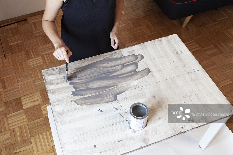 从上方看，穿黑色衬衫的女性正在家中将旧白色木桌涂成灰色。图片素材