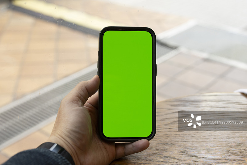 亚洲人手持黑色智能手机，屏幕为绿色，背景模糊的咖啡店内。图片素材