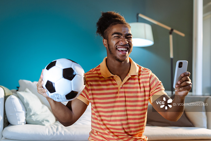 兴奋的年轻男子在家中观看足球比赛，手里拿着智能手机和足球。图片素材