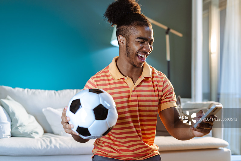 兴奋的年轻人在家里室内拿着足球和智能手机图片素材