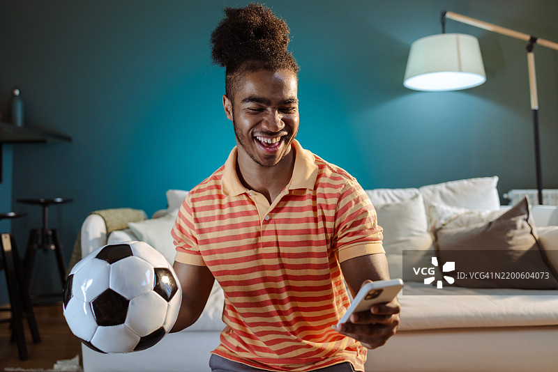 微笑的男人在舒适的客厅里拿着足球并浏览手机图片素材