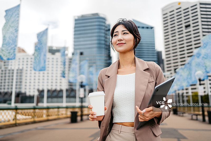 年轻的亚洲女商人在澳大利亚悉尼达令港一边喝外带咖啡，一边拿着笔记本电脑。图片素材