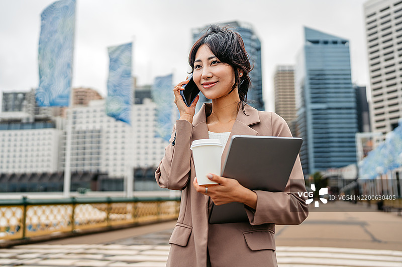 年轻的亚洲女商人在澳大利亚悉尼的达令港一边打电话一边喝外带咖啡，同时手里还拿着一台笔记本电脑。图片素材