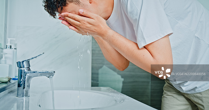 年轻男子在家浴室的镜子前用洁面乳洗脸。健康生活方式的早晨日常概念。图片素材