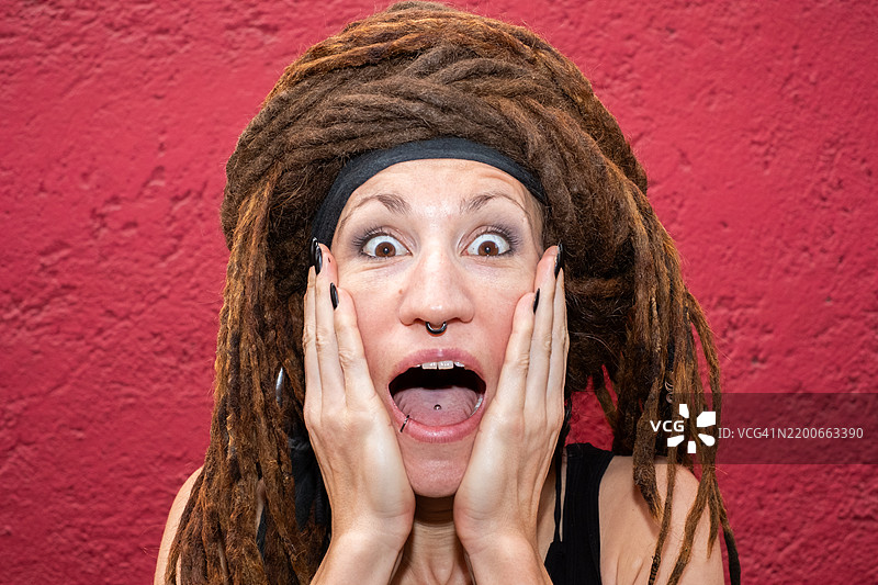 一位戴着头巾的白人女性，头发是脏辫，惊讶地用手捂住脸看着镜头，表情震惊。图片素材