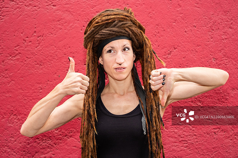 一位戴着头巾的白人女性，头发是脏辫，困惑地看着镜头，同时做出竖起大拇指和竖下大拇指的手势。图片素材