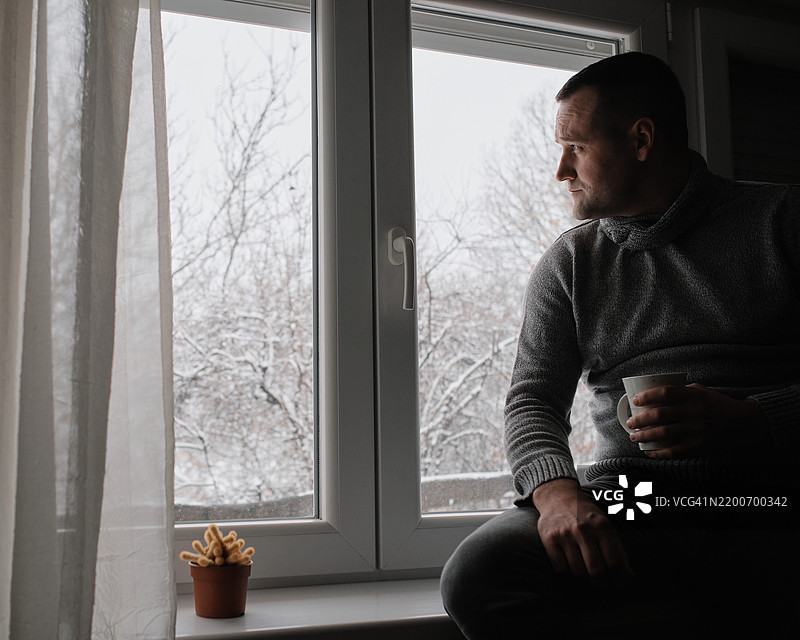 一位年轻帅气的男子坐在窗边，喝着早晨的咖啡。窗外是一幅冬季田园风光。图片素材