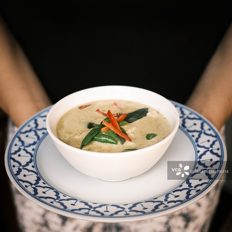 泰式椰子鸡汤（冬阴功）特写，配有辣椒和新鲜香草，盛在白色碗中，放在传统的蓝白陶瓷盘上，手持于黑色背景前，展现正宗泰国美食和食品摄影概念图片素材