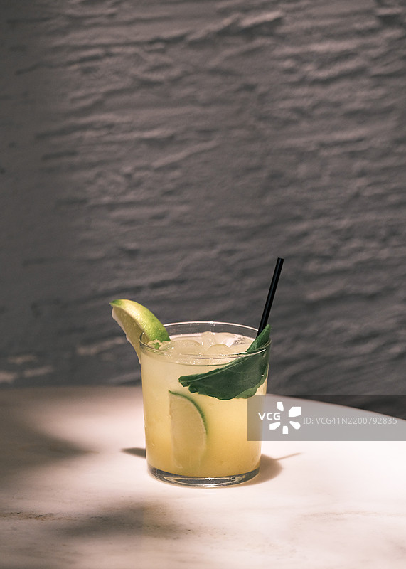 清新的青柠鸡尾酒，配冰块、罗勒装饰和黑色吸管，盛在短玻璃杯中，摆放在白色桌子上，背景是有纹理的灰色墙壁，优雅的调酒和夏季饮品概念图片素材