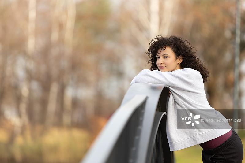一个美丽的女孩，留着深色卷发，正在公园里放松，微笑着四处张望。图片素材