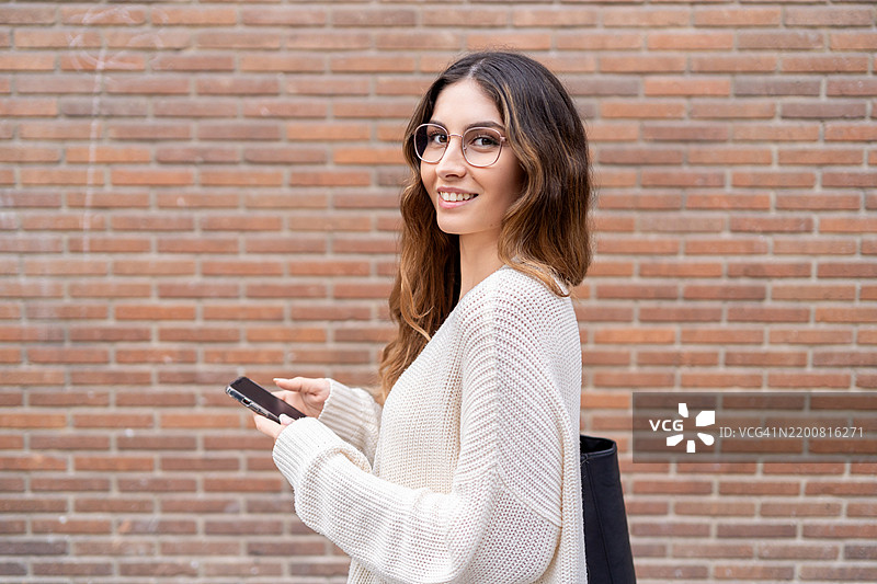 年轻女性戴着眼镜，自信地站在砖墙旁，轻松地使用智能手机。图片素材