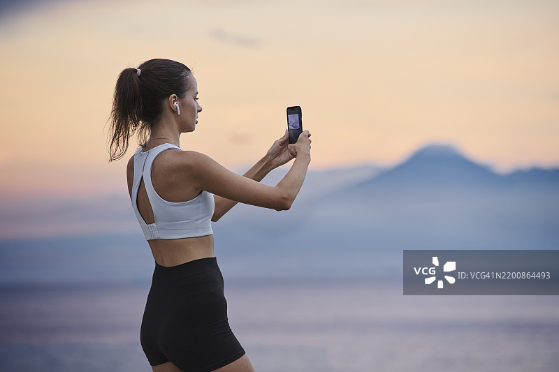 年轻女性穿着运动服，使用手机拍摄日落时海洋和阿贡火山的景色图片素材