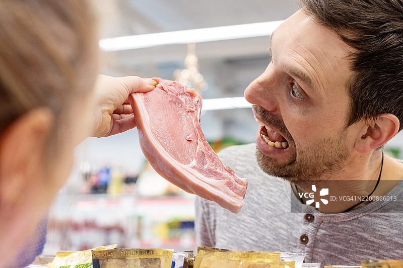 顾客在肉铺柜台假装咬生猪排。超市里有趣的购物时刻图片素材