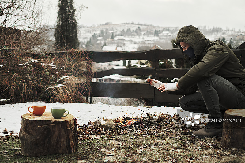 一位英俊的年轻男子穿着冬季服装，在山坡上生火露营，四周被雪和寒冷包围。图片素材