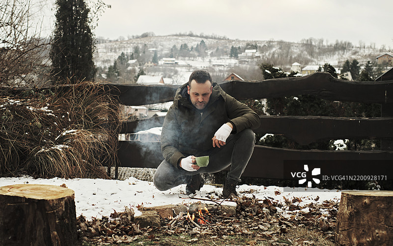 一位英俊的年轻男子穿着冬季服装，在山坡上生火露营，四周被雪和寒冷包围。图片素材