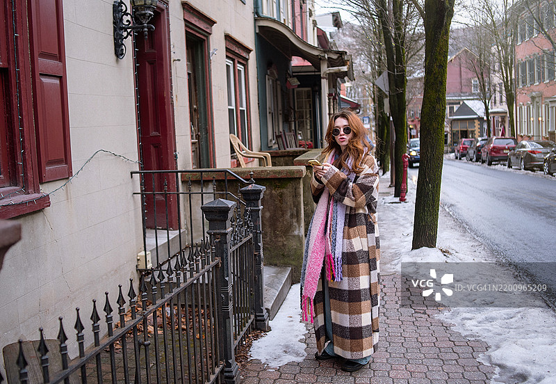 千禧一代在宾夕法尼亚小镇捕捉冬季回忆。时尚的24岁年轻人在覆盖着雪的吉姆·索普维多利亚区穿着格子外套和粉色围巾探索。图片素材