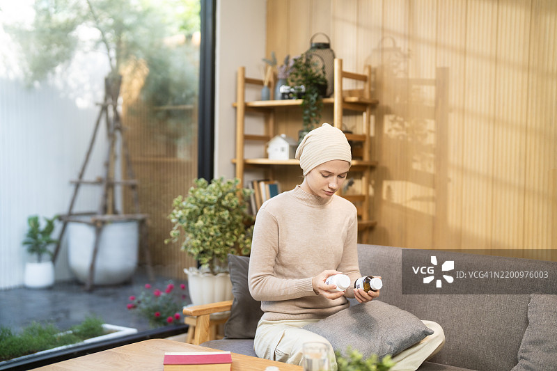 一位成年癌症患者戴着头巾，坐在客厅的沙发上，手里拿着两瓶药物并在检查。温馨的氛围，配有自然装饰和家具。图片素材