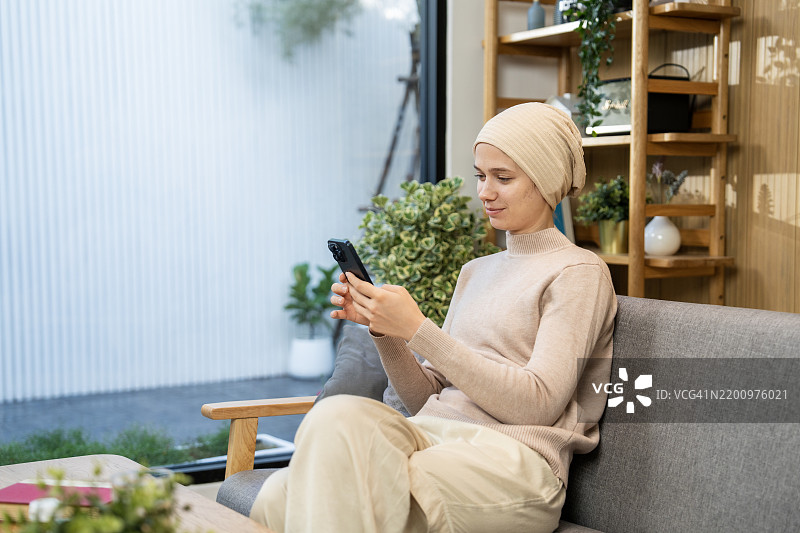 女性癌症患者坐在室内，使用智能手机，身处一个舒适现代明亮的房间。图片素材