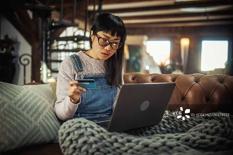 年轻的日本女性在客厅使用信用卡在线购物，身边有智能手机和笔记本电脑。图片素材