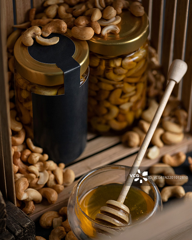 玻璃罐中的腰果和蜂蜜，搭配细长勺子。蜂蜜甜点。天然自制的甜味。健康饮食，无糖。食品概念，随时可食用。小吃。美味的礼物。留白空间。特写。图片素材