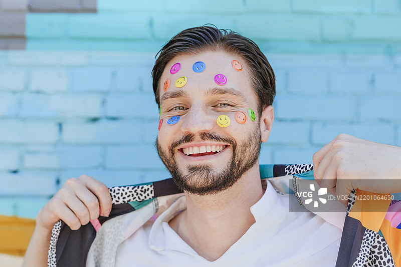 面带微笑的男子在砖墙前用手捏着衣领，脸上贴着贴纸。图片素材