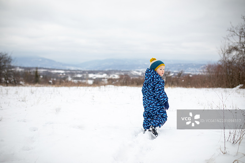 可爱的小男孩肖像，穿着冬季衣物，站在被雪覆盖的草地上，背景是美丽的城市景观。图片素材