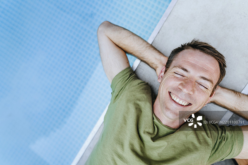 快乐的男人躺在游泳池旁，双手放在头后。图片素材