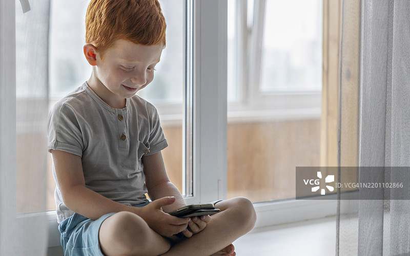 红发男孩坐在窗台上，盘腿使用智能手机图片素材