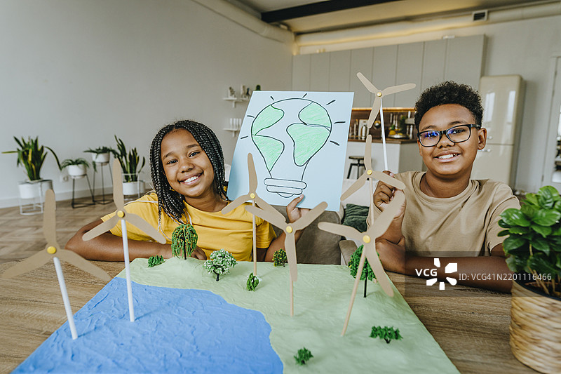 快乐的兄弟姐妹坐在桌子旁，进行风力发电机项目和灯泡绘画。图片素材